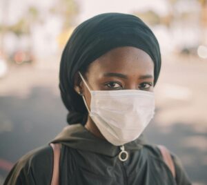 Unrecognizable young ethnic woman in jacket and headscarf and protective mask standing on city street
